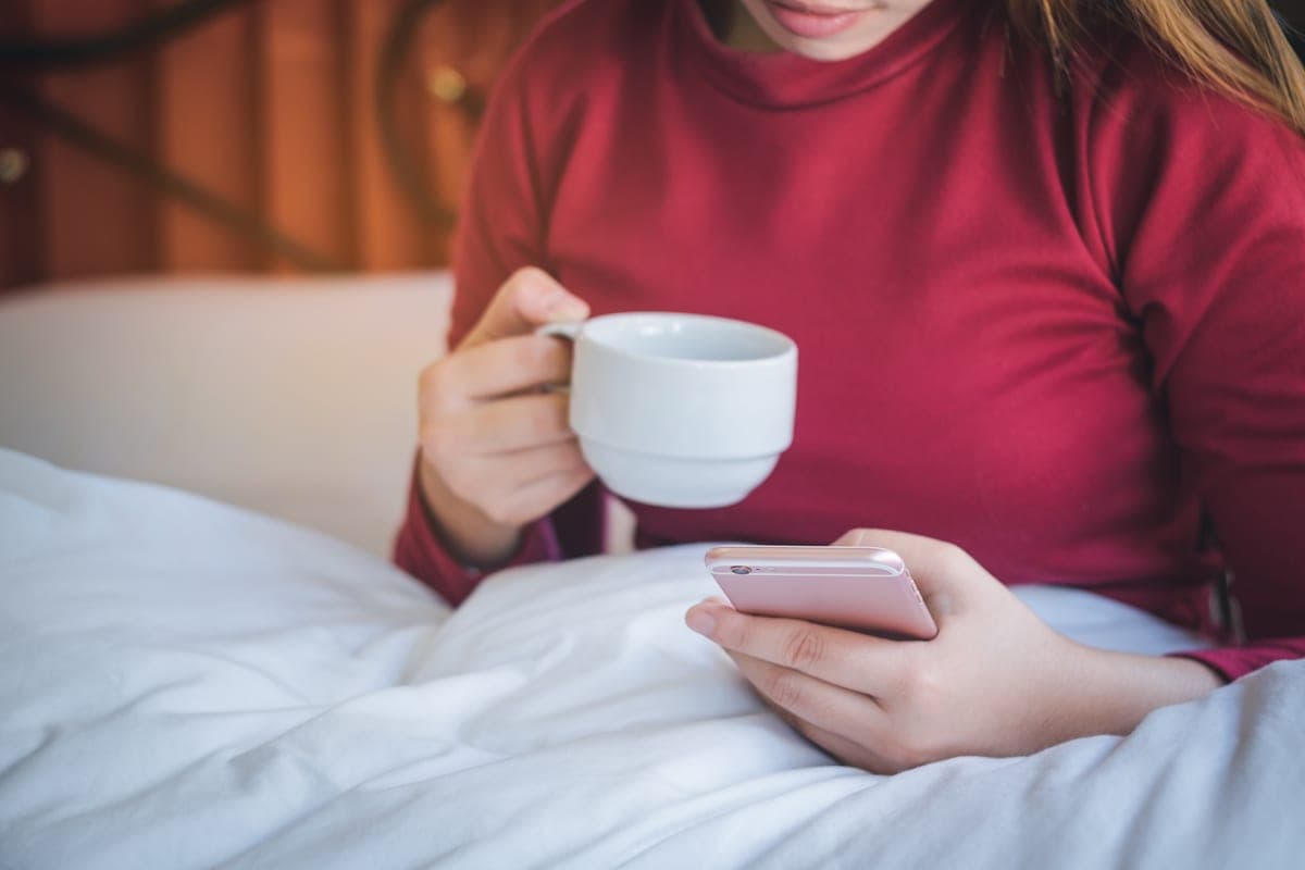 Žena v posteli pod peřinou s hrnkem v ruce píše na telefonu