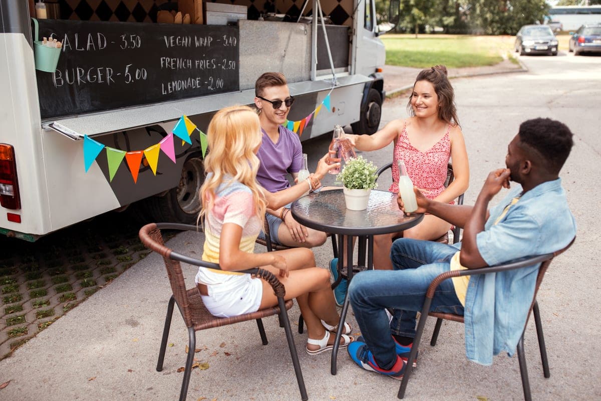 Přátelé sedí u stolku před food truckem s tabulí s nabídkou jídel