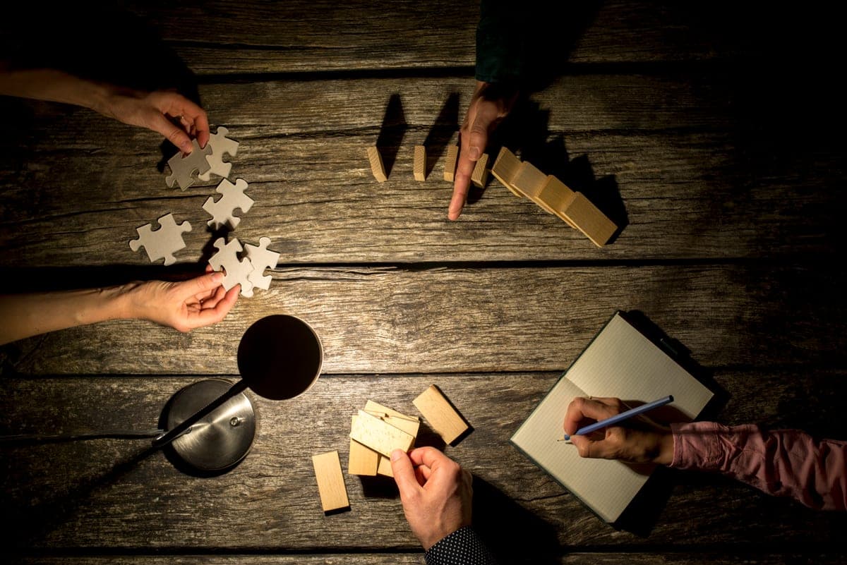 Ruce několika lidí společně skládají puzzle a dřevěné kostky na tmavém stole